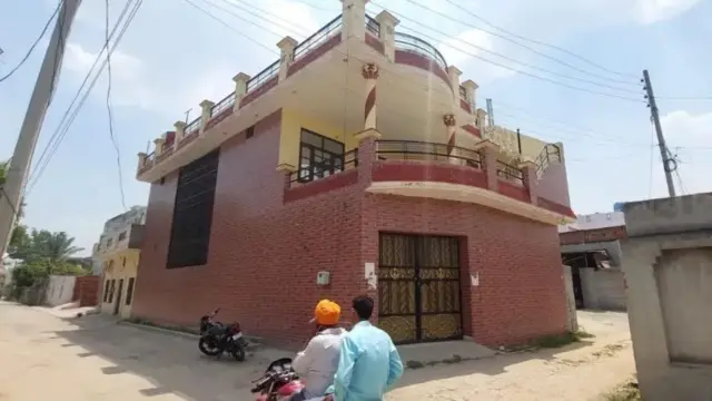 ਘਰ ਦੇ ਬਾਹਰ ਦੀ ਤਸਵੀਰ , ਘਰ ਅੱਗਿਓਂ ਮੋਟਰਸਾਈਕਲ ਸਵਾਰ ਦੋ ਜਣੇ ਲੰਘਦੇ ਹੋਏ।