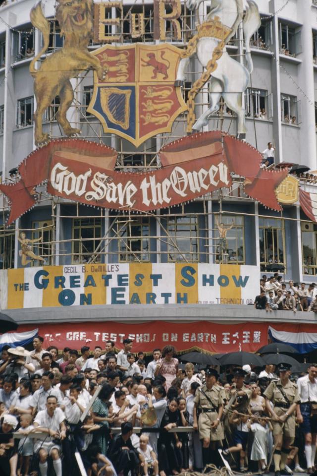 香港九龙旺角弥敦道上市民在“天佑女王”大型牌匾下围观庆祝英女王登基巡游（3/6/1953）