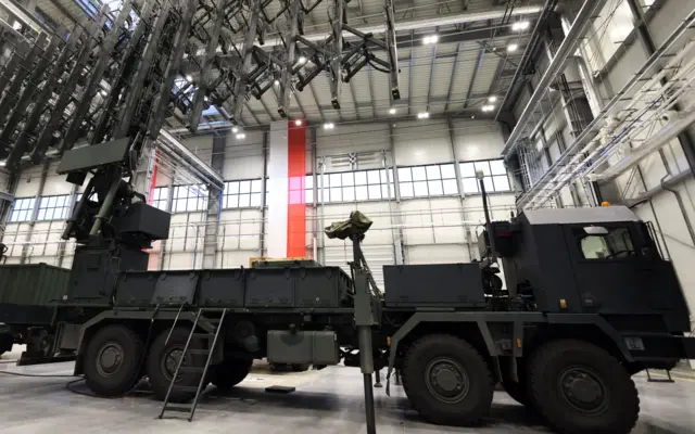 Sprzęt wojskowy ustawiony wewnątrz dużej hali serwisowej, z widocznym pojazdem ciężarowym wyposażonym w rozbudowaną konstrukcję antenową