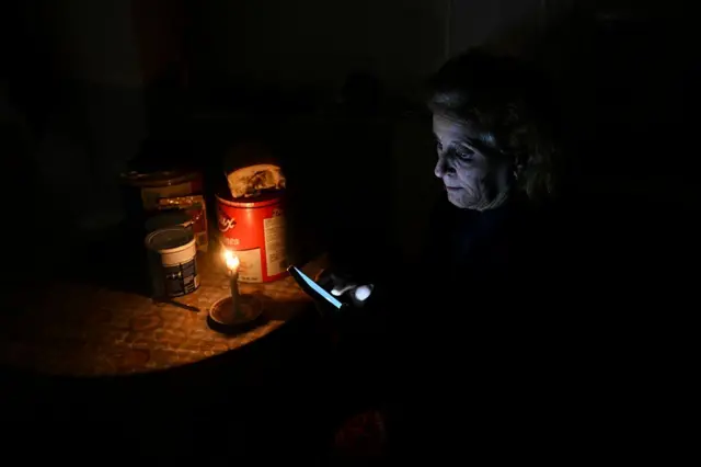Una mujer sentada junto a una mesa con su rostro iluminado por una vela y el celular que sostiene en sus manos.