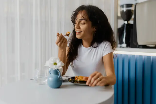 امرأة شابة تجلس على طاولة وتأكل من طبق باستخدام شوكة، وهي مغمضة العينين ومستمتعة بالطعام. تظهر في الخلفية ستائر بيضاء وخلفية زرقاء.