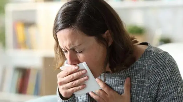 Жінка у сірій кофті кашляє, прикладаючи хустинку до вуст