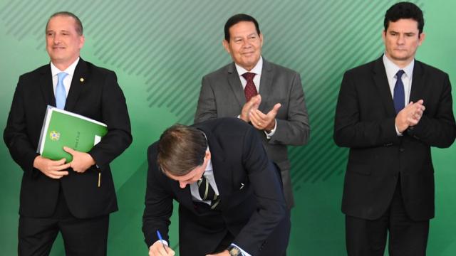 Jair Bolsonaro assinando documento ao lado dos ministros Onyx Lorenzoni, Sergio Moro e do vice-presidente Hamilton Mourão