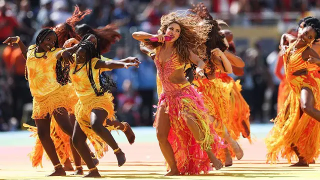 Shakira, pembe, sarı, turuncu ve kırmızı renklerde uçuşan bir kıyafetle, arka plandaki dansçılar eşliğinde dans ediyor.