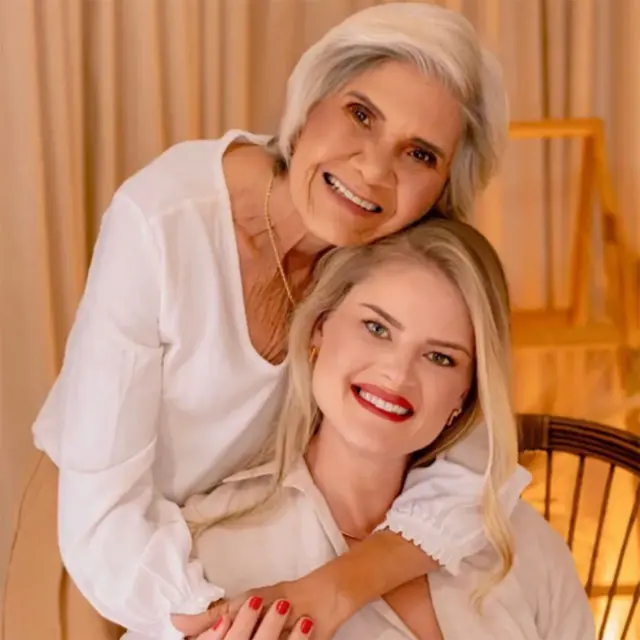 一位留着金色长发的年轻女子坐在椅子上。一位留着齐下巴灰发的年长女子站在她身后，双臂环抱着她的肩膀。她们都穿着白色上衣，面带微笑。