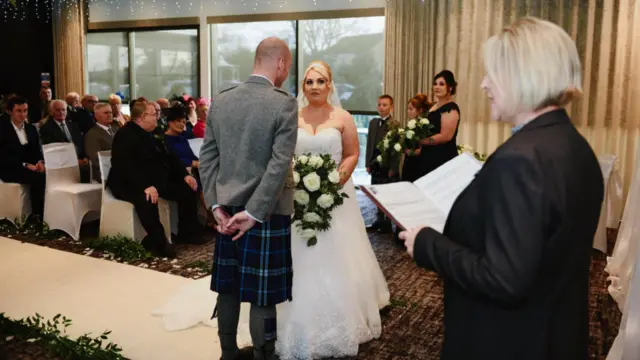 Uma cerimônia de casamento, com os noivos frente a frente e os convidados assistindo.