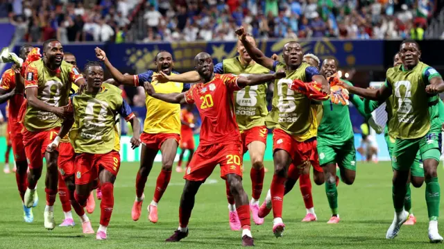 Yoane Wissa étend les bras en dansant pour célébrer après avoir marqué un but contre l'Ouzbékistan et lors de la finale de la Coupe du monde. Il est entouré de plusieurs remplaçants de la RD Congo, qui arborent tous le sourire alors que certains d'entre eux copient sa pose
