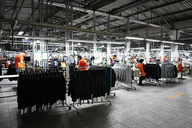 Kıyafetlerin askılarla durduğu bir atölyede işçiler çalışıyor. 