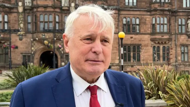 George Duggins se encuentra frente a la sede del Ayuntamiento de Coventry y viste un traje azul y una corbata roja.