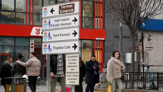 Yalova sokaklarından bir görüntü. Binalar, tabelalar ve yürüyen insanlar görülüyor. 