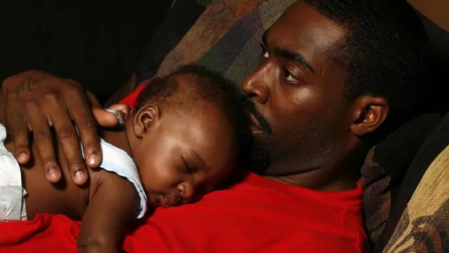 رجل يرتدي قميصاً أحمر اللون ويحمل طفلاً نائماً على صدره