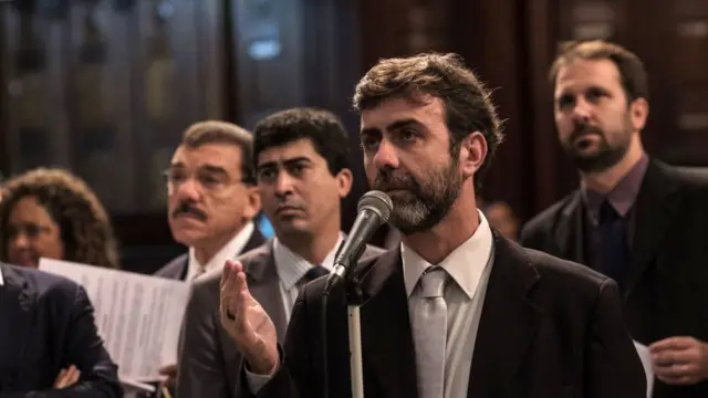 Marcelo Freixo falando no microfone em assembleia, rodeado por outras pessoas com trajes formais