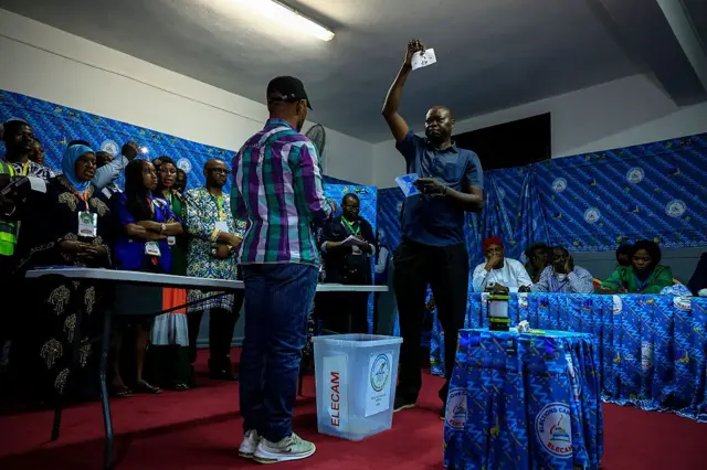 Des observateurs électoraux (à gauche) regardent un responsable électoral tenir un bulletin de vote de Paul Biya, président du Cameroun et candidat à la présidence pour le Rassemblement des démocrates du Cameroun (RDPC), pendant le dépouillement des votes au bureau de vote de l'école publique Bastos à Yaoundé, le 12 octobre 2025,