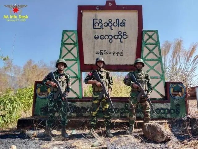 ကြိုဆိုပါ၏ မကွေးတိုင်းလို့ ရေးထားတဲ့ မြို့အဝင်ဆိုင်းဘုတ်အရှေ့မှာ သေနတ်ကိုင်ပြီး မတ်တပ်ရပ်နေတဲ့ စစ်ဝတ်စုံပြည့်နဲ့ အေအေတပ်ဖွဲ့ဝင် ၃ ဦး 