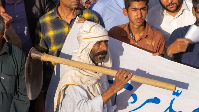 Beyaz bir örtü takmış ve cübbe giymiş bir adam, diğer protestocular yanında dururken omzunda bir çapa tutuyor