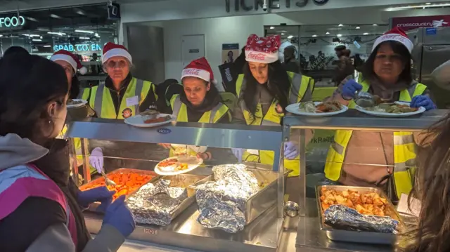 Cinco mujeres con chalecos reflectantes y gorros de Papá Noel sirven comida a personas sin hogar.