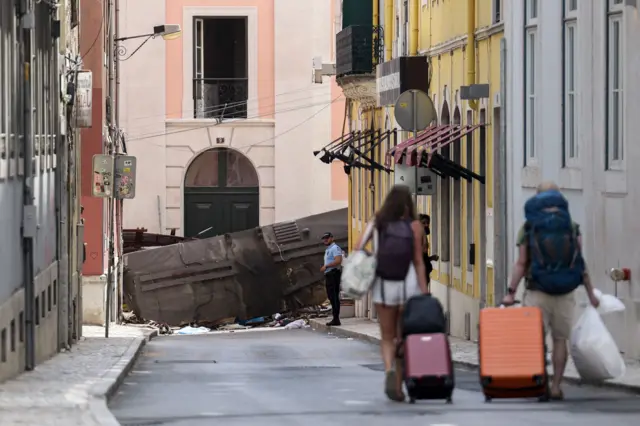 Duas mulheres de costas carregam malas em rua estreita, com bondinho caído ao fundo