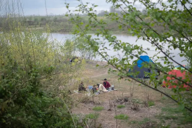یک خانواده روی فرشی در کنار رودخانه نشسته‌اند در حالی که پشت‌آنها دو چادر دیده می‌شود