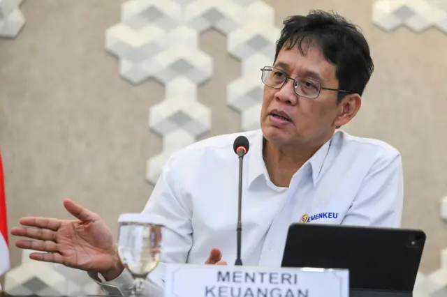 Menteri Keuangan Purbaya Yudhi Sadewa menggunakan baju putih polos dengan tulisan Kemenkeu. Ia nampak sedang bicara kepada orang di hadapannya.