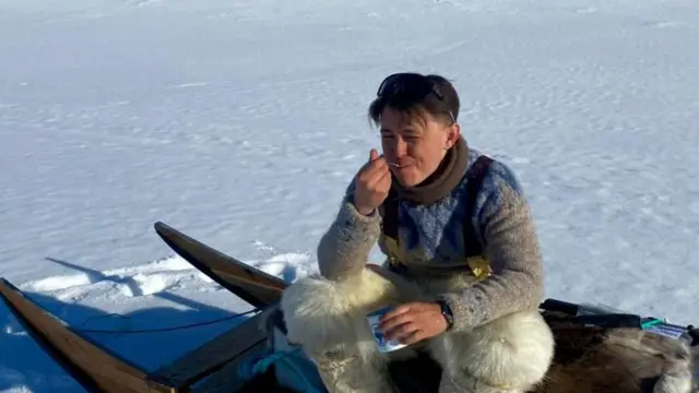 Un hombre sentado sobre un trineo en una superficie helada come con una cuchara. 