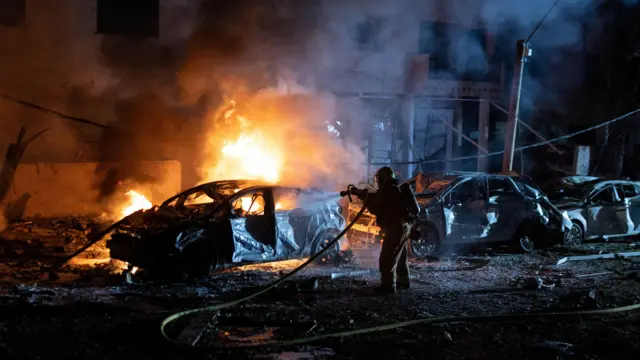 A firefighter at night projects water from a hose at a burning car that is lit up in orange flames; other cars around it are severely damaged works.
Picture from Itai Ron / Reuters - no access to Israeli outlets