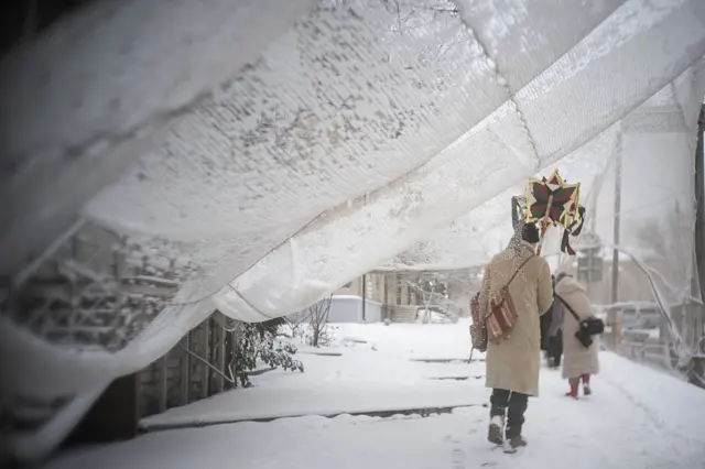 Зимова вулиця, вкрита снігом. Двоє людей у теплих пальтах ідуть спиною до камери; один несе різдвяну зірку над головою. Над доріжкою провисає біла сітка, усе навколо вкрите снігом.