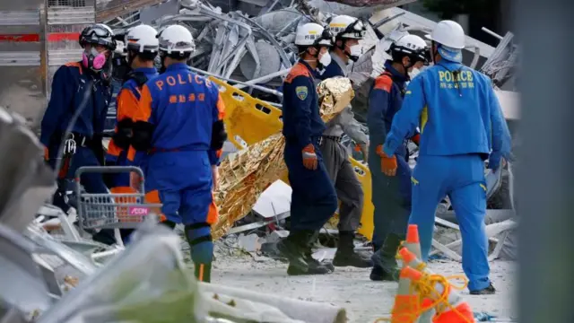 がれきの散乱する中、さまざまな熊本県警などさまざまな所属の隊服を着た人たちたヘルメットやマスクをつけて立っている