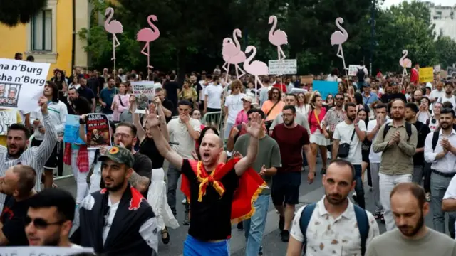 Un numeroso grupo de personas, algunas gritando, otras con los brazos en alto y coreando, participa en una manifestación. Algunas sostienen siluetas de flamencos rosas, el símbolo de la protesta.