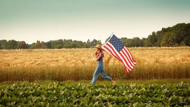 Farmer girl carrying an American flag marching across a field of crops