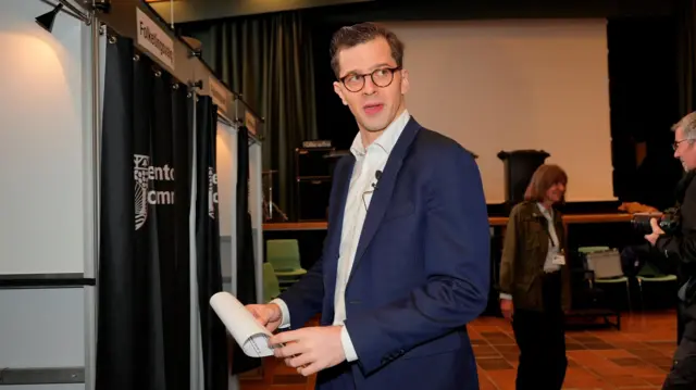 Молодой мужчина в очках и в костюме перед избирательной кабинкой, в руках у него лист бумаги.