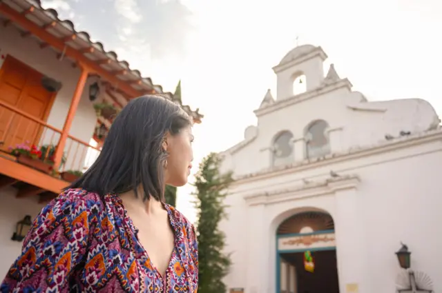 Młoda kobieta o czarnych włosach, w sukience w kolorowe wzory, oddala wzrok od obiektywu, patrząc na zabytkowy kościół o bielonych ścianach. Cerro Nutibara, Kolumbia. 