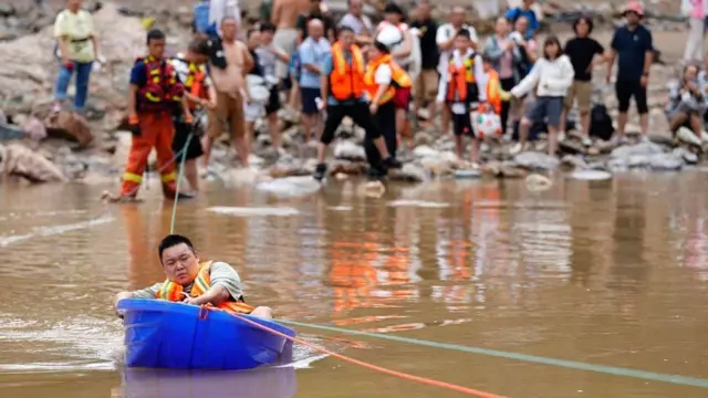 青色の大きなプラスチック容器に、髪の短い、オレンジ色のベストを着けた人が乗り、ロープに沿うように水面を移動している。背後の水辺では救急隊員や多くの人たちが立って見ている