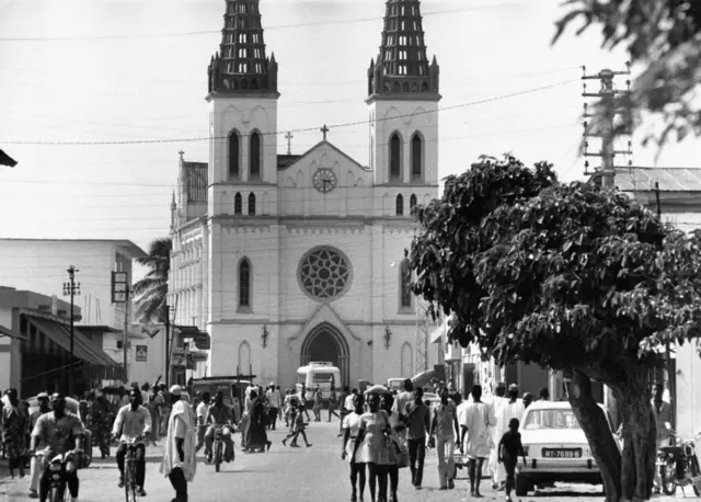 Des gens marchent sur une rue de Lomé à l'époque coloniale avec le bâtiment de la cathédrale visible au fond.