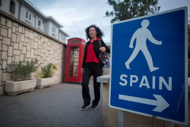 Mujer caminando al lado de una típica cabina de teléfonos británica de color rojo y, al otro lado y en primer plano, un cartel que indica que se puede ir caminando a "Spain", "España".