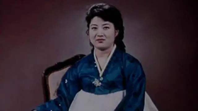 Ko Yong-hui, provavelmente na casa dos 30 ou 40 anos, usa um vestido coreano azul e branco e sorri para a câmera.