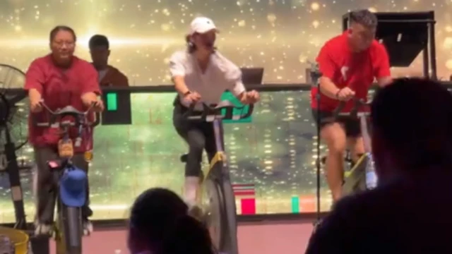 Na scenie, oświetlonej jasnymi światłami, trzy osoby ćwiczą na rowerach stacjonarnych. Widać sylwetki dwóch uczestników jadących na rowerach i zwróconych w stronę instruktorów.