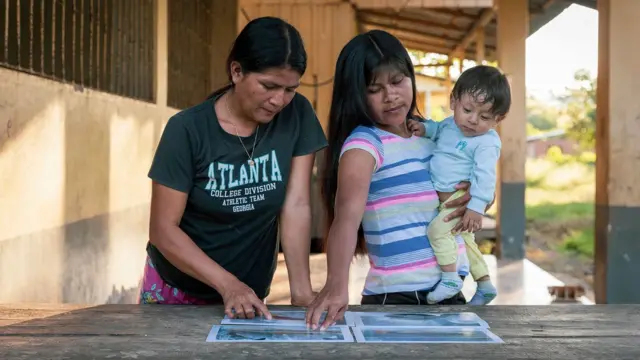Dos mujeres indigenas en Ecuador señalan fotos de peces. Una de ellas tiene un bebé en brazos. 