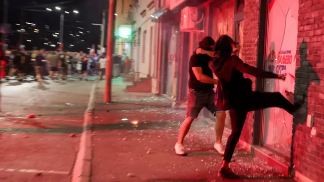 Участники акции протеста темной одежде и масках ногами бьют стекла в офисе правящей партии. На заднем фоне видна толпа демонстрантов