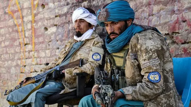 Pakistan'la sınır kapısında nöbet tutan Afgan Taliban milisi iki adam, bir duvarın önünde birer sandalye üzerinde yan yana oturmuş. İkisi de yorgun gözüküyor ve yere doğru bakıyorlar. Üstlerinde askeri kamuflaj kıyafetleri, başlarında birer geleneksel örtü, ellerinde ise birer tüfek var.