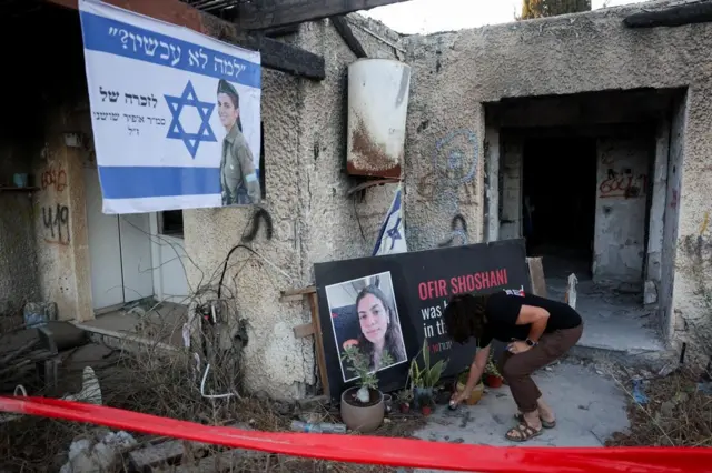 женщина зажигает свечу у плаката с портретом женщины в разрушенном доме в кибуце Кфар-Аза
