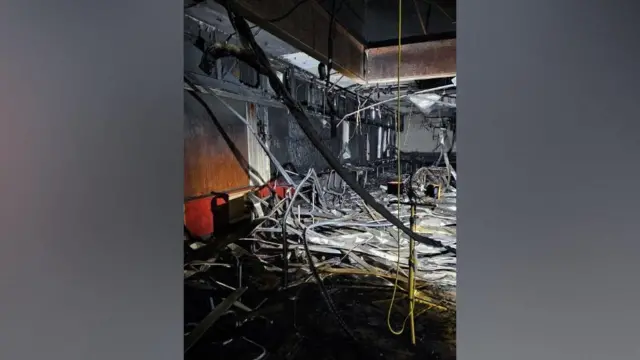 Una habitación con paredes de paneles ennegrecidos, tuberías de metal colgando del techo y muebles esparcidos por el suelo.