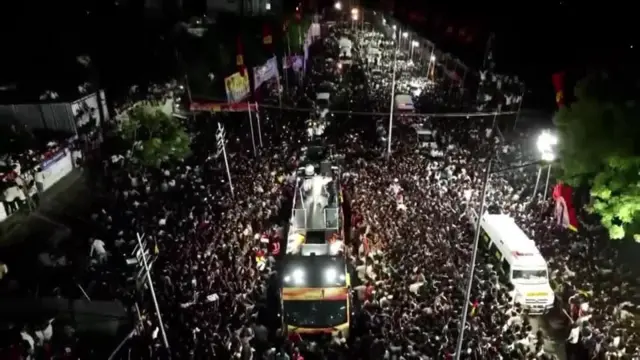 도로 근처에 많은 군중이 모여 있고, 흰 옷을 입은 작은 인물이 차량 위에 서 있는 모습이 보인다.