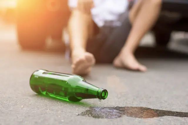 drop of beer dripping from a tilted beer bottle on road, a collapsed man in background.