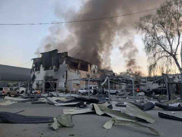 На фото видно сильно пошкоджену будівлю з обгорілими стінами, з якої підіймається густий чорний дим. Навколо розкидані уламки металевих конструкцій, поруч стоять пошкоджені автомобілі, місцями видно осередки вогню.