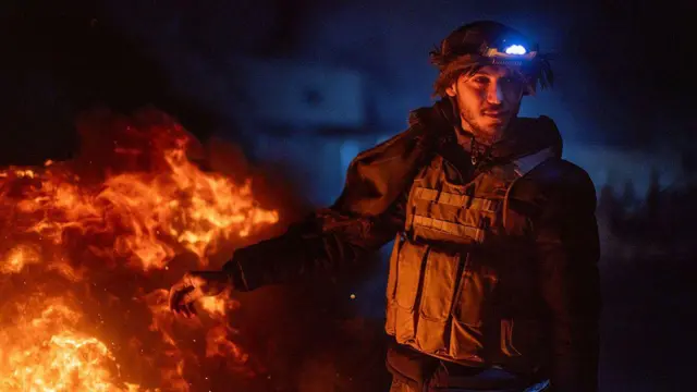 Kamışlı'da nöbet tutan bir Kürt savaşçı. Yanında ateş yanıyor. Üzerinde çelik yelek ve uzun namlulu silah bulunuyor.