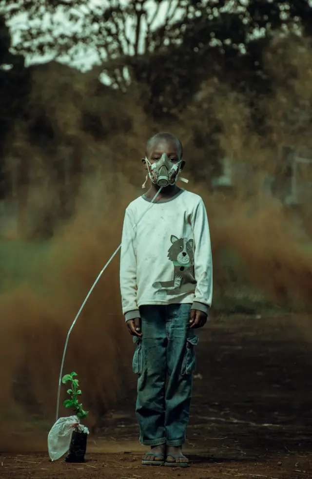 A boy is seen wearing an oxygen mask attached to a plant, with a sand storm behind him