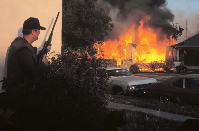 Un agente del FBI tiene una escopeta en las manos, mientras en el fondo se ve una casa incendiándose.