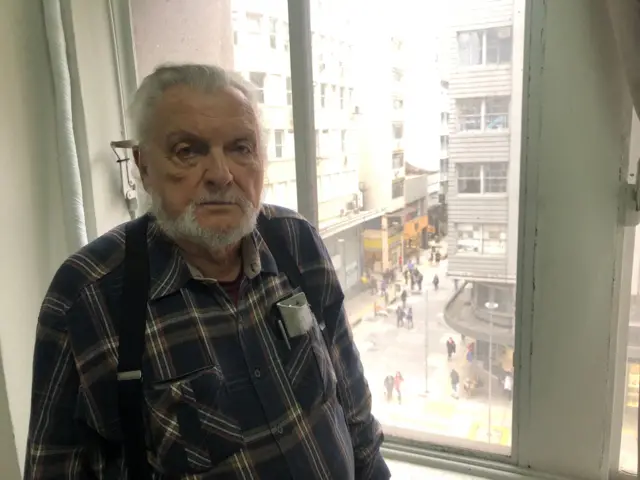 Jair Krischke, um homem idoso de cabelos e barba branca, vestindo camisa xadrez, olha para a câmera com expressão séria, ao lado de uma janela com vista para a rua
