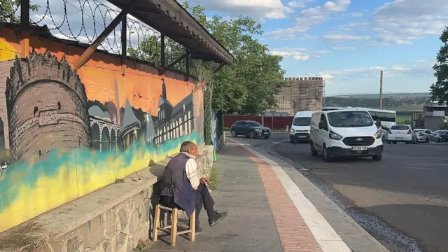 Diyarbakır'da bir sokakta kaldırıma oturmuş adamın elinde sigara var. Arabaların geçişini izliyor gibi. Arkasında Diyarbakır surlarının resmi bir duvara çizilmiş