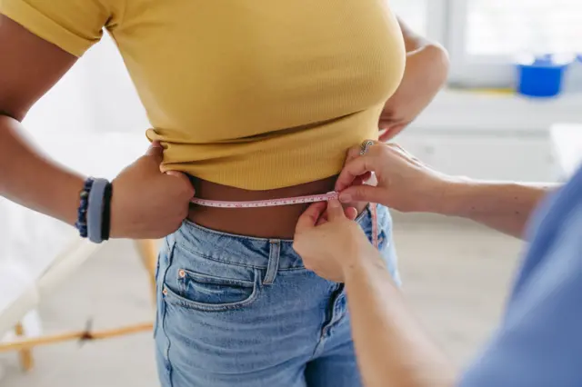 A woman lifts up her yellow top as another woman wraps a tape measure around her waist